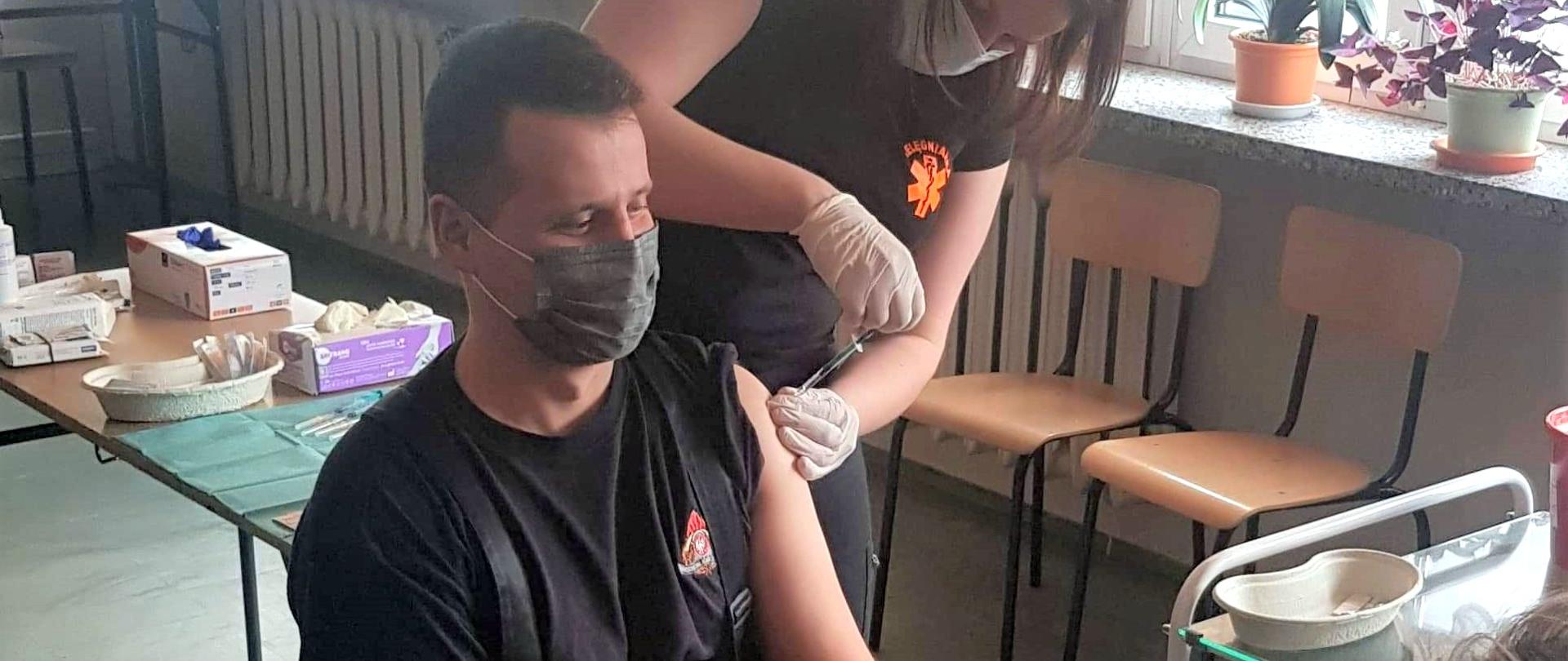 Strażak w umundurowaniu dowódczo sztabowym w czarnym podkoszulku przyjmuje szczepionkę