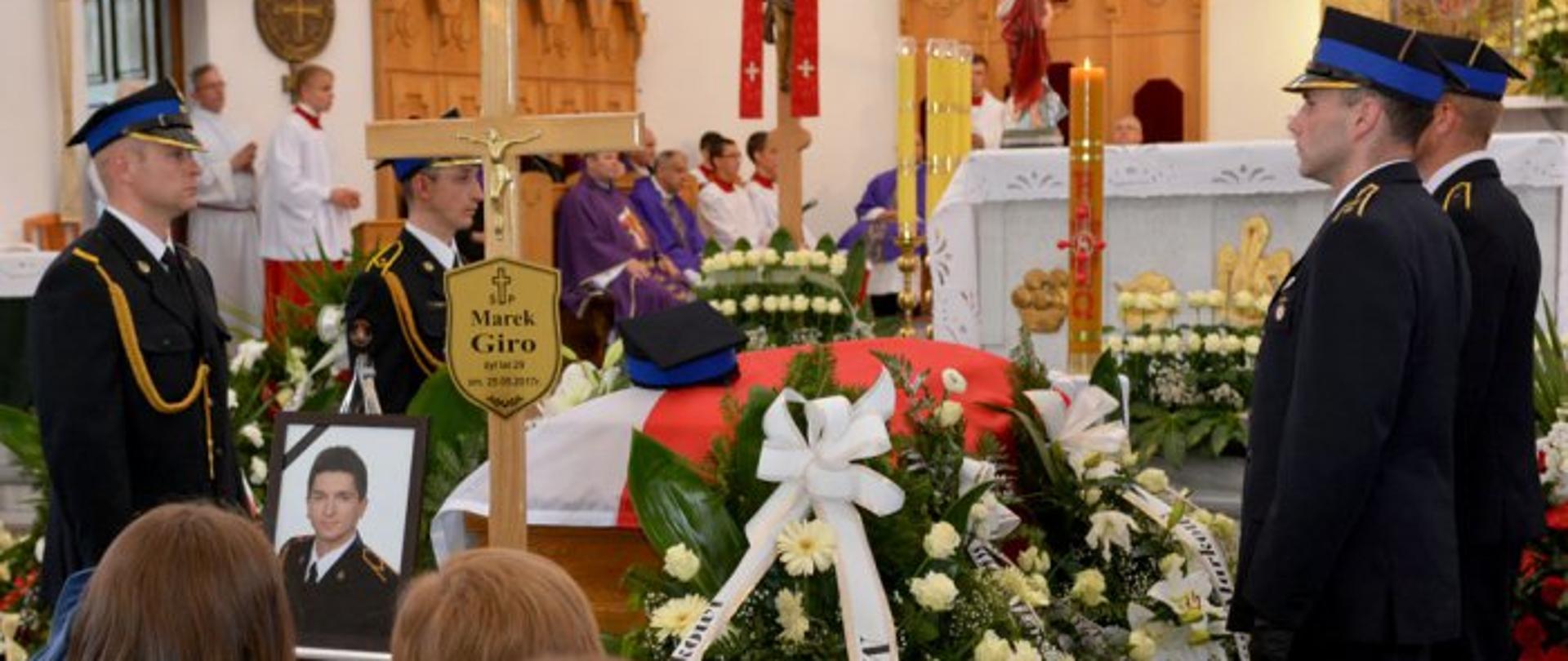 Trumna nakryta flagą biało - czerwoną, po obu stronach stoją strażacy , przed trumną ustawiona fotografia zmarłego strażaka