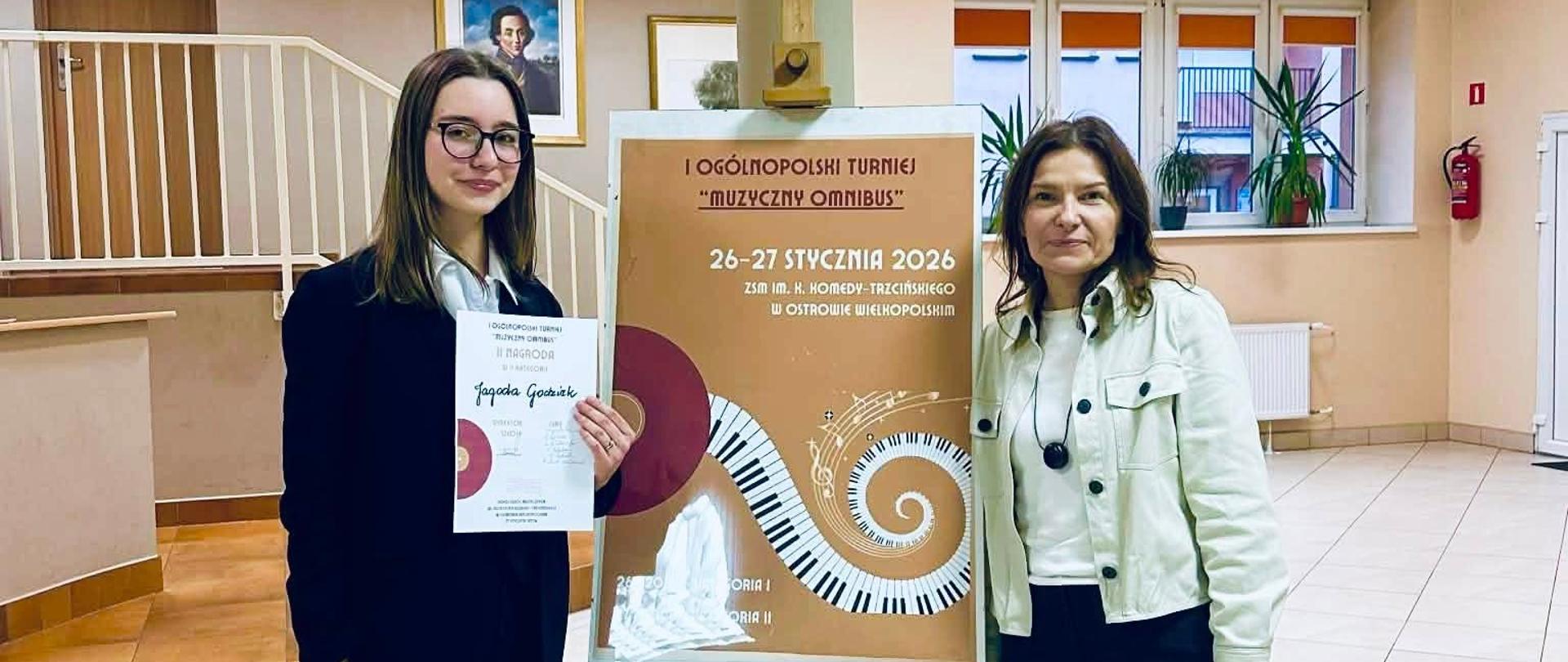 Na zdjęciu znajdują się dwie kobiety stojące w jasnym wnętrzu budynku szkoły. Po lewej stronie Jagoda Godziek młoda kobieta w okularach trzyma dyplom i papierową torbę, a po prawej Dorota Kwiatoń-Strzelecka nauczycielka uśmiecha się do aparatu. Między nimi stoi sztaluga z plakatem informującym o wydarzeniu „I Ogólnopolskie Turnieje Muzyczny Omnibus”, który odbył się 26–27 stycznia 2026 roku w Sokołowie Podlaskim.