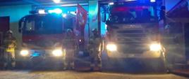 Widoczni strażacy na tle pojazdów gaśniczych i garaży oddający hołd zmarłym strażakom