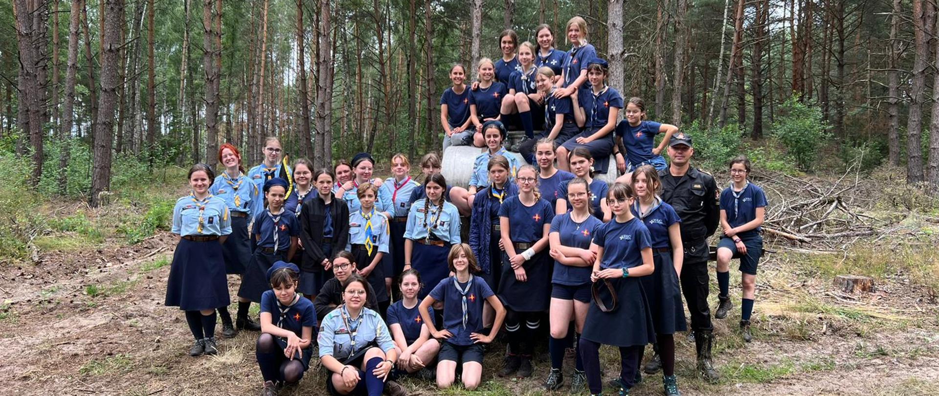 Wizytacja obozu harcerskiego w Rytelach Święckich - na zdjęciu w lesie na polanie harcerze w strojach harcerskich stoją w grupie do zdjęcia, z nimi strażak w ubraniu sztabowym.