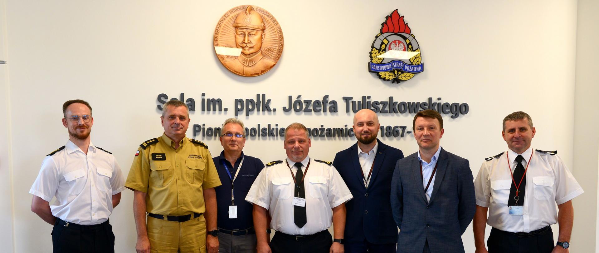 W szeregu ustawieni członkowie SITP wraz z Komendantem Wojewódzkim PSP woj. pomorskiego. W tle ściana z napisem "Sala imienia podpółkownika Józefa Tuliszkowskiego - pioniera polskiego pożarnictwa.
