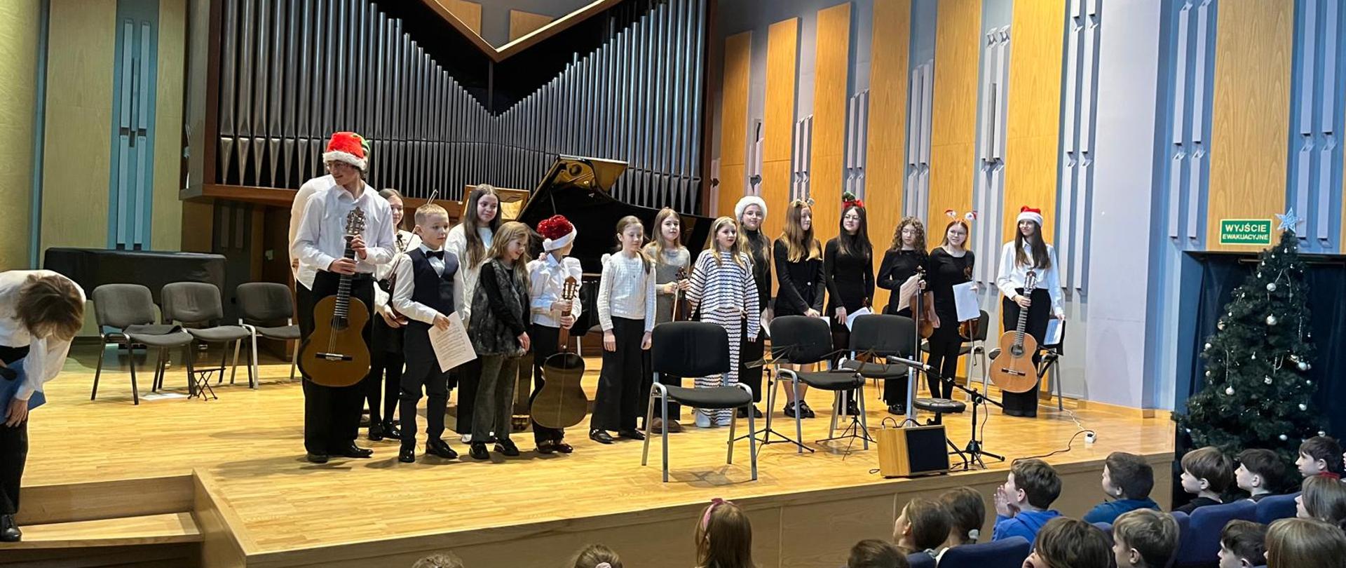 Zdjęcie przedstawia grupę dzieci i młodzieży na scenie podczas koncertu bożonarodzeniowego w sali szkoły muzycznej. Wykonawcy: Na drewnianym podwyższeniu stoi kilkanaścioro młodych artystów. Niektórzy trzymają gitary klasyczne, inni mają w rękach nuty. Część osób ma na głowach czapki św. Mikołaja lub świąteczne opaski. Są ubrani galowo (białe koszule, ciemne spodnie) lub w stroje czarno-białe. Instrumenty i wyposażenie: W tle dominuje ogromny, nowoczesny prospekt organowy o metalowych piszczałkach ułożonych w kształt litery "V". Obok widać otwarty czarny fortepian skrzydłowy. Na scenie ustawione są również czarne krzesła dla muzyków oraz mikrofony. Publiczność: W dolnej części zdjęcia widać tyły głów dzieci siedzących w rzędach niebieskich foteli, które obserwują występ Otoczenie: Po prawej stronie stoi przystrojona choinka. Ściany sali są wyłożone jasnymi panelami akustycznymi. W prawym górnym rogu widać zielony napis „WYJŚCIE EWAKUACYJNE”. Atmosfera zdjęcia jest uroczysta i radosna, sugeruje zakończenie występu lub przygotowania do wspólnego utworu.
