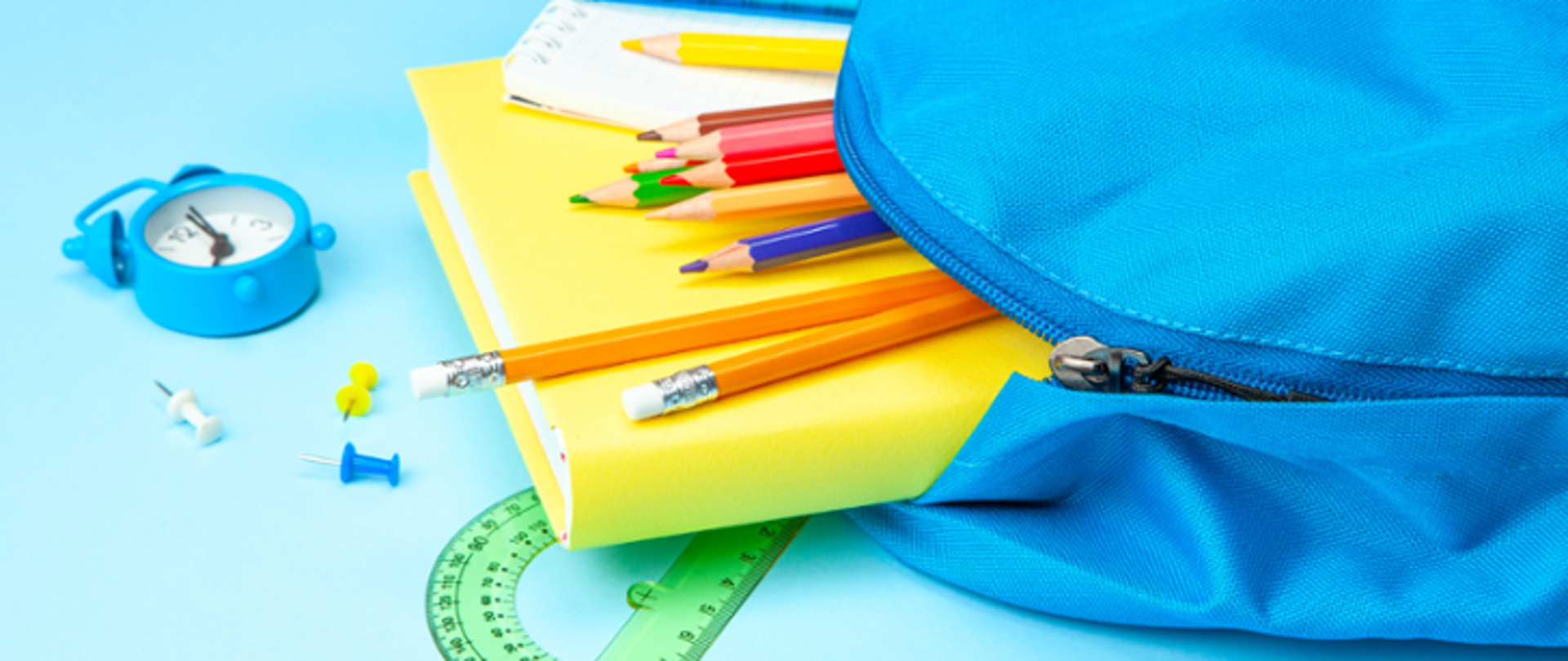 Na zdjęciu rozsunięty niebieski plecak, na jasnoniebieskim tle rozsypane przybory szkolne (kredki, ołówki, kątomierz), z plecaka wysuwa się żółty podręcznik i notes.
