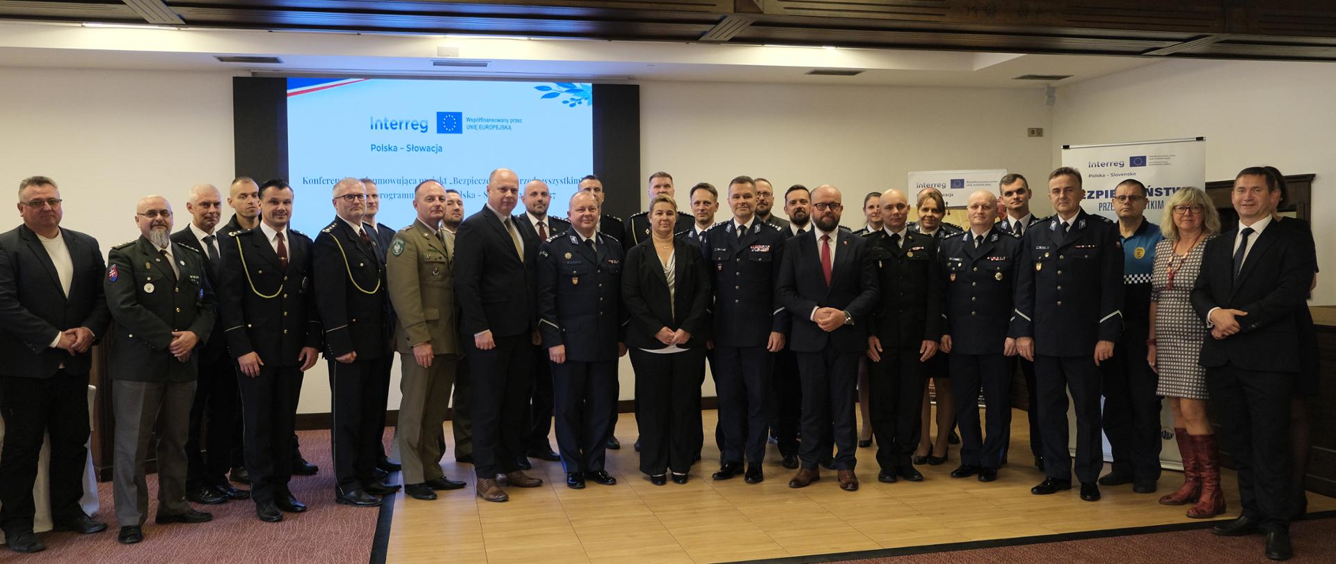 Na zdjęciu widać kilkadziesiąt osób, uczestników spotkania, stoją w dwóch rzędach na tle ekranu z logo Interreg Polska – Słowacja 2021-2027