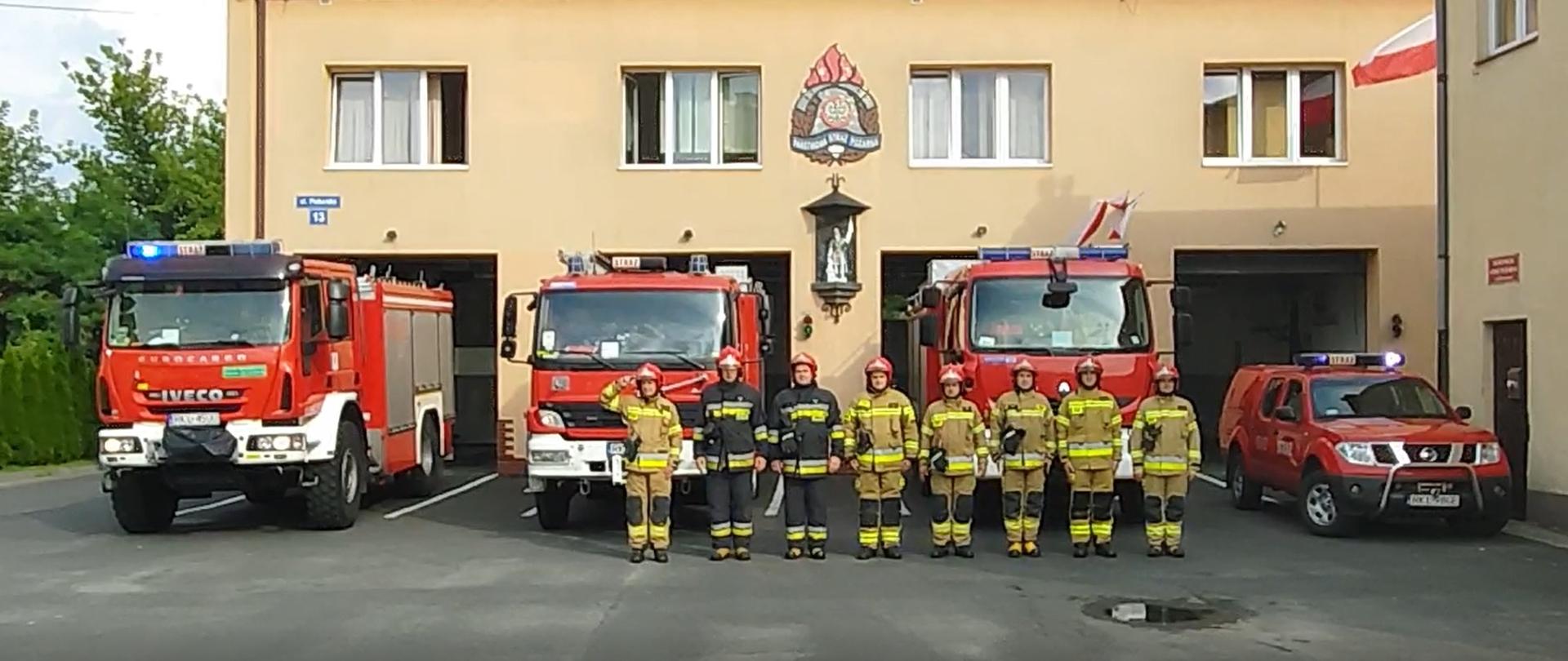 Na zdjęciu cztery samochody strażackie widziane z przodu z włączonymi światłami alarmowymi: trzy ciężarowe i jeden osobowy. Przed nimi stoi 8 strażaków w ubraniach bojowych, 6 ubrań w kolorze piaskowym, 2 w czarnym. Jeden ze strażaków salutuje. W tle budynek Jednostki Ratowniczo - Gaśniczej w Kolbuszowej.