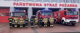 Zdjęcie przedstawia dziewięciu strażaków stojących w pozycji zasadniczej przed budynkiem Komendy Powiatowej PSP w Łowiczu. Jeden strażak po lewej stronie zdjęcia salutuje, za nimi stoją trzy pojazdy straży pożarnej z włączonymi sygnałami błyskowymi, wszyscy są ubrani w ubrania bojowe typu Moratex z odblaskami barwy szarej i żółtej, na głowie mają hełm a na twarzy maseczkę chirurgiczną. W tle budynek komendy oraz napis na froncie PAŃSTWOWA STRAŻ POŻARNA.