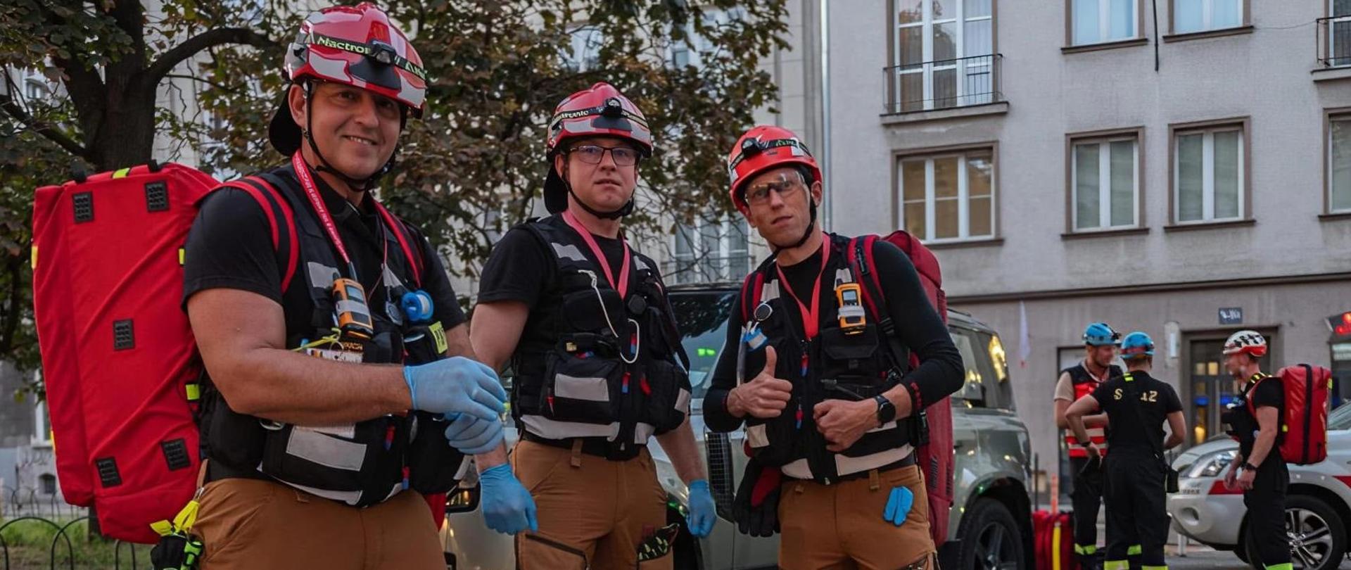 Trzej strażacy w ratowniczych kaskach, taktycznych kamizelkach i brązowych spodniach stoją na ulicy przed budynkiem. Jeden z nich pokazuje kciuk w górę. 