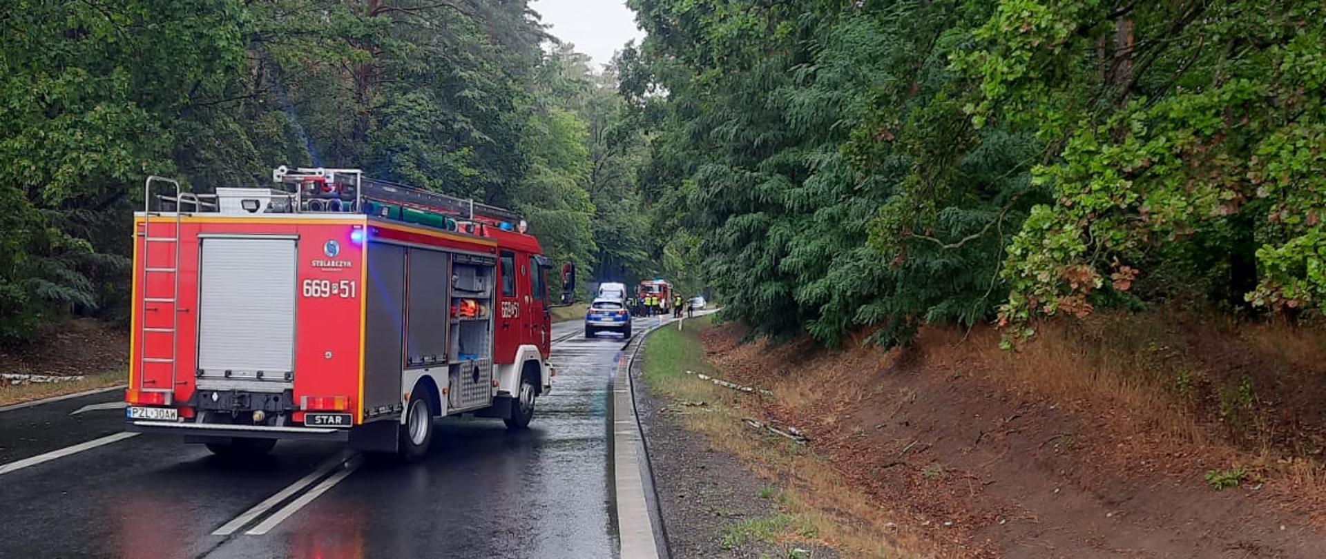 Na zdjęciu droga asfaltowa mokra czarna świecąca na ,której stoi samochód strażacki