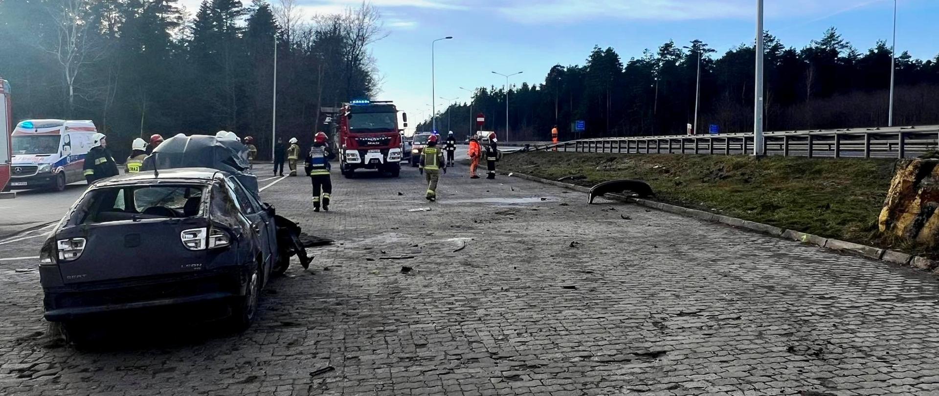 Samochód osobowy po dachowaniu stoi z uszkodzeniami na kołach. Niedaleko niego chodzą strażacy. W tle widać samochód strażacki i karetkę pogotowania ratunkowego. 