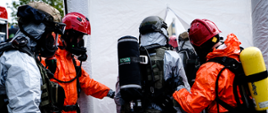 Policjanci oraz strażacy w ubraniach chemicznych i aparatach ochrony dróg oddechowych stoją przed namiotem dekontaminacyjnym.