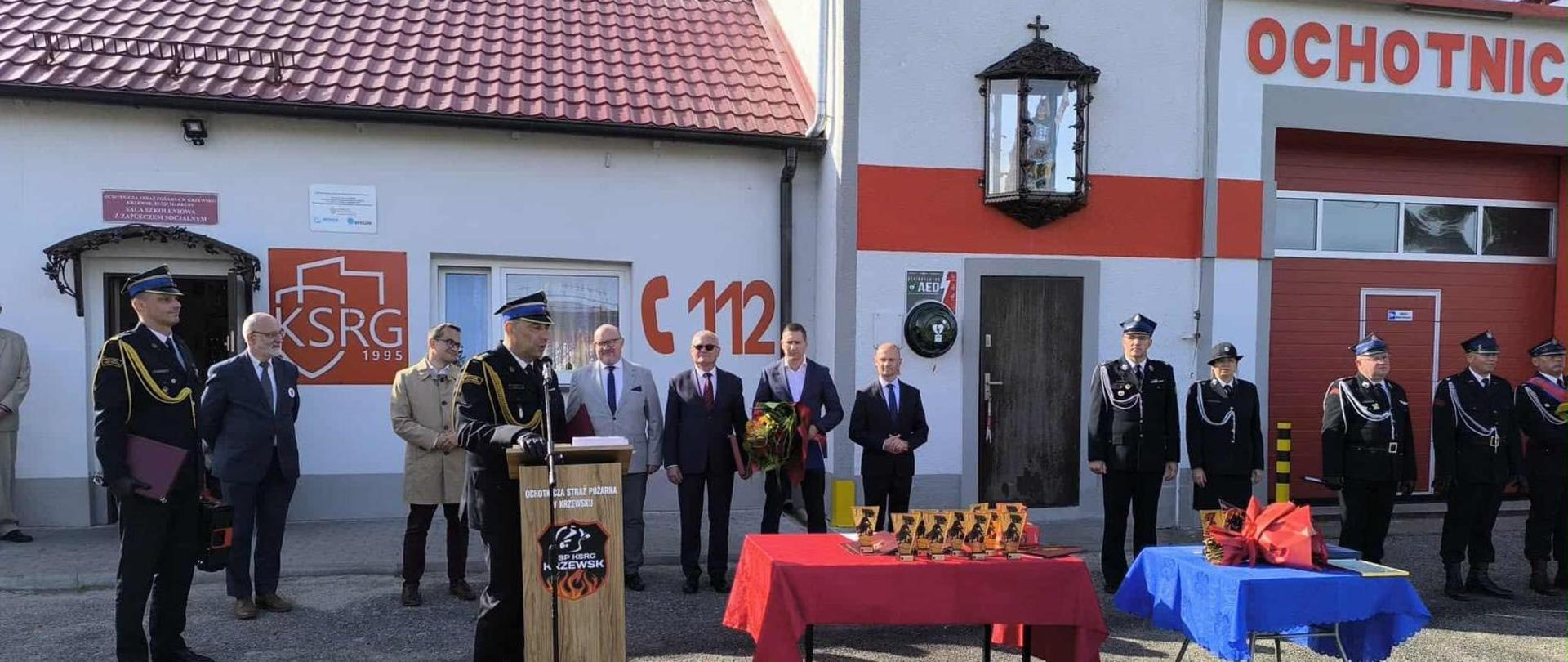 Funkcjonariusze państwowej straży pożarnej, druhowie ochotniczych straży pożarnych oraz zaproszenie goście znajdują się na placu przed remizą strażacką. Jeden z funkcjonariuszy stoi za mównicą. 