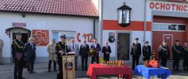 Funkcjonariusze państwowej straży pożarnej, druhowie ochotniczych straży pożarnych oraz zaproszenie goście znajdują się na placu przed remizą strażacką. Jeden z funkcjonariuszy stoi za mównicą. 