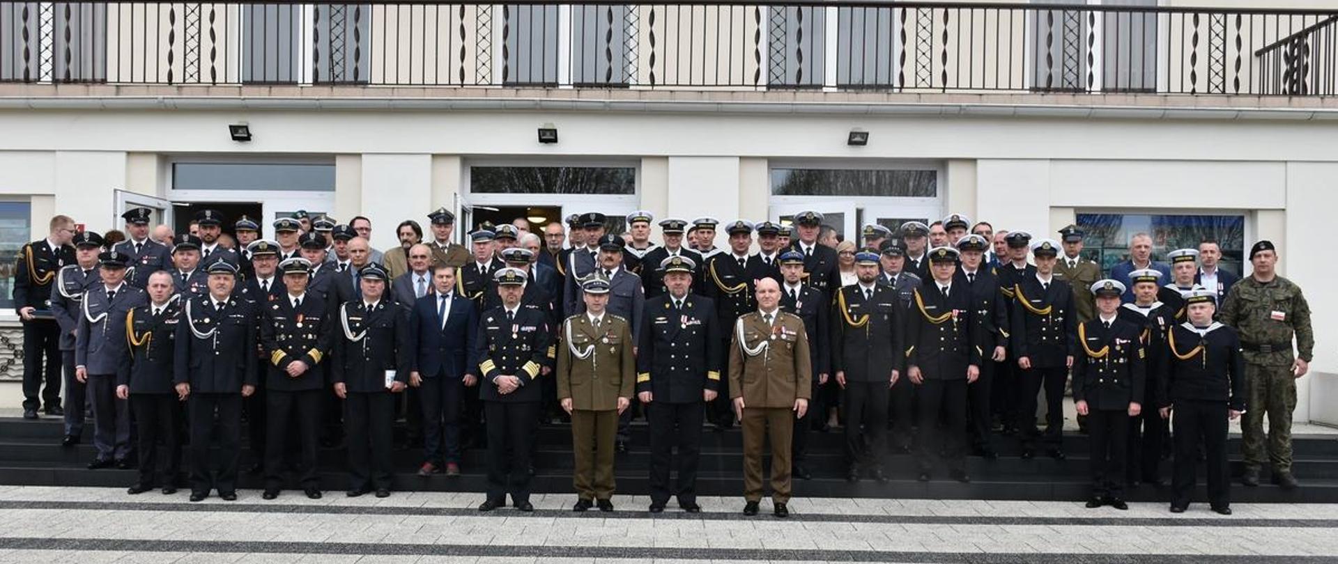 Żołnierze strażacy w mundurach wyjściowych ze sznurem stoją obok siebie przed wejściem do budynku.