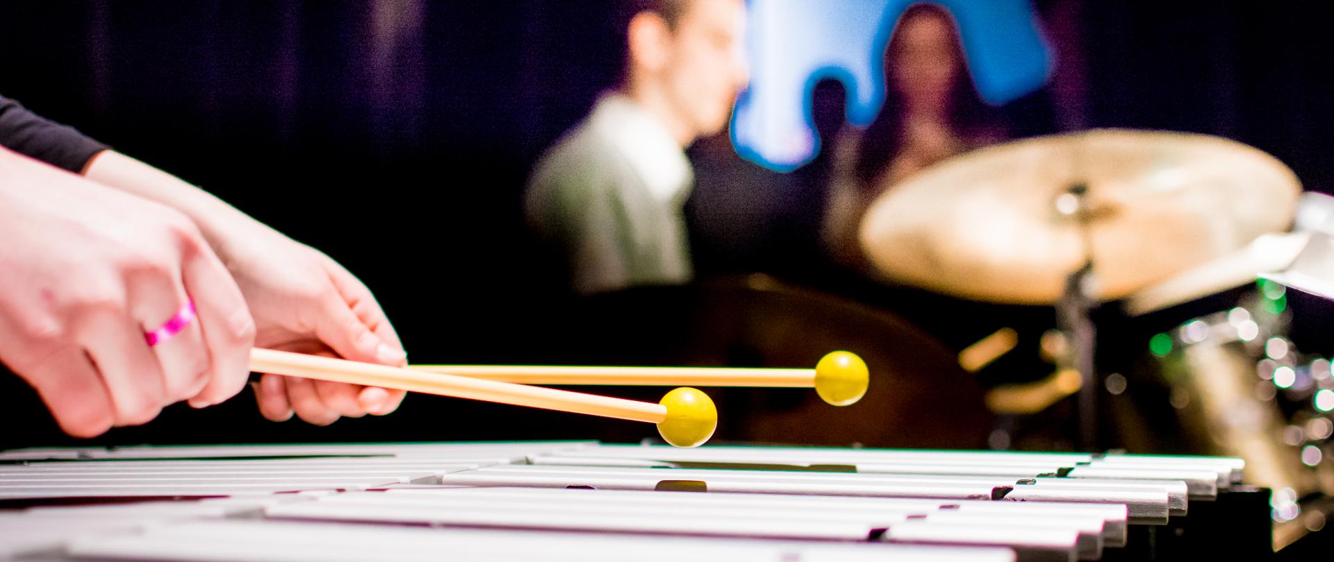 Ręce ucznia grającego na wibrafonie, w tle uczeń grający na zestawie perkusyjnym