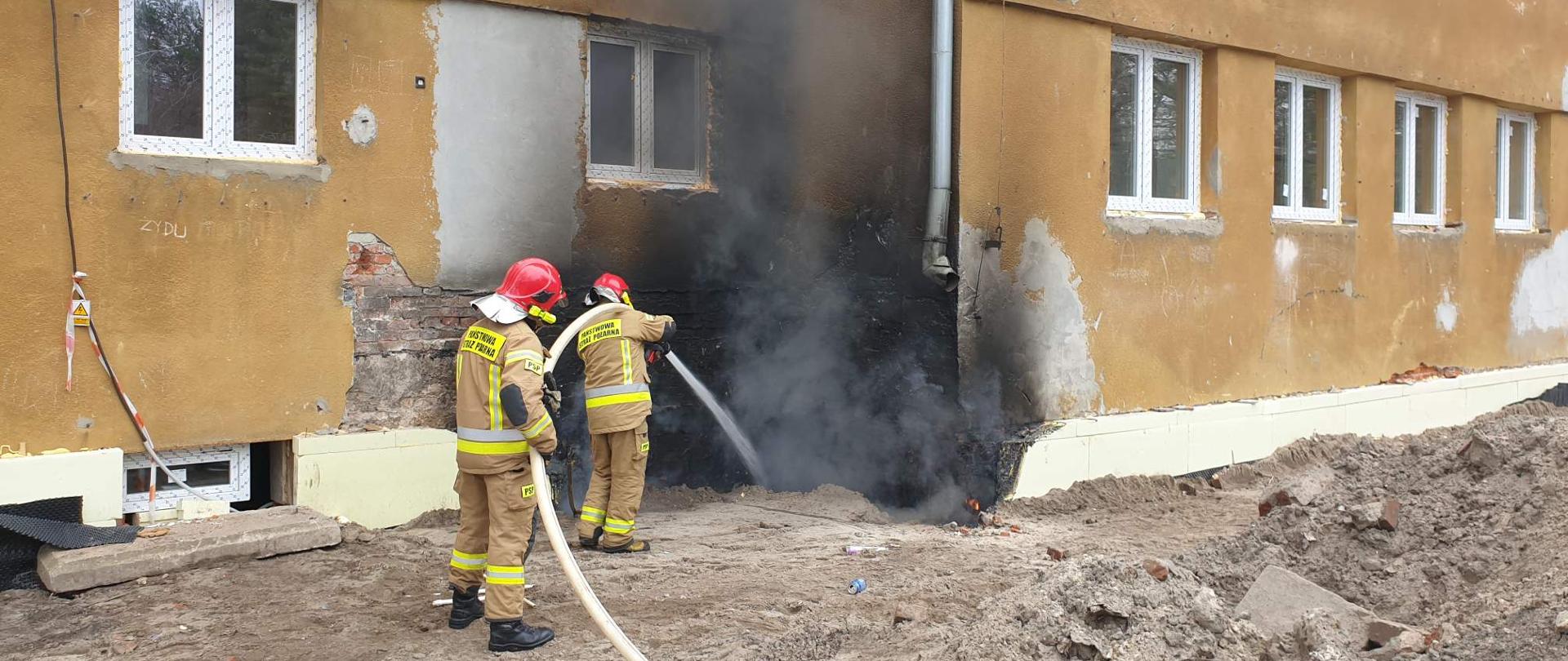 Dwóch strażaków trzyma węża gaśniczego, którym podawana jest woda. Woda skierowana jest na elewację budynku, z której wydostają się kłęby dymy.
gasi wodą podawana wężem gasniczympalący się styropian, 