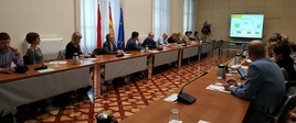 Konsultacje Pełnomocników ds. otwartości danych z instytucjami hiszpańskimi. Ludzie siedzą przy owalnym stole. W tle flagi Polski, UE i Hiszpanii.