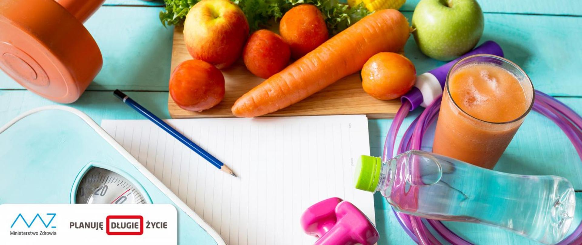Leżące na stole: ciężarki, skakanka, waga, ołówek, kartka papieru, warzywa, owoce, butelka wody, szklanka soku.