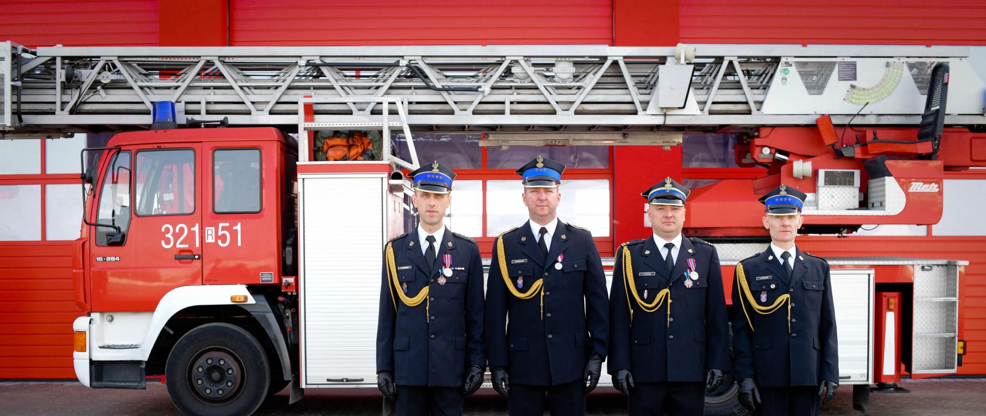 Zdjęcie przedstawia sylwetki czterech strażaków odchodzących na zaopatrzenie emerytalne, ubranych w mundury wyjściowe w postawie zasadniczej. Strażacy stoją na tle wozu - drabiny strażackiej wystawionej przed garaż Jednostki Ratowniczo-Gaśniczej Nr 3 w Rzeszowie.
