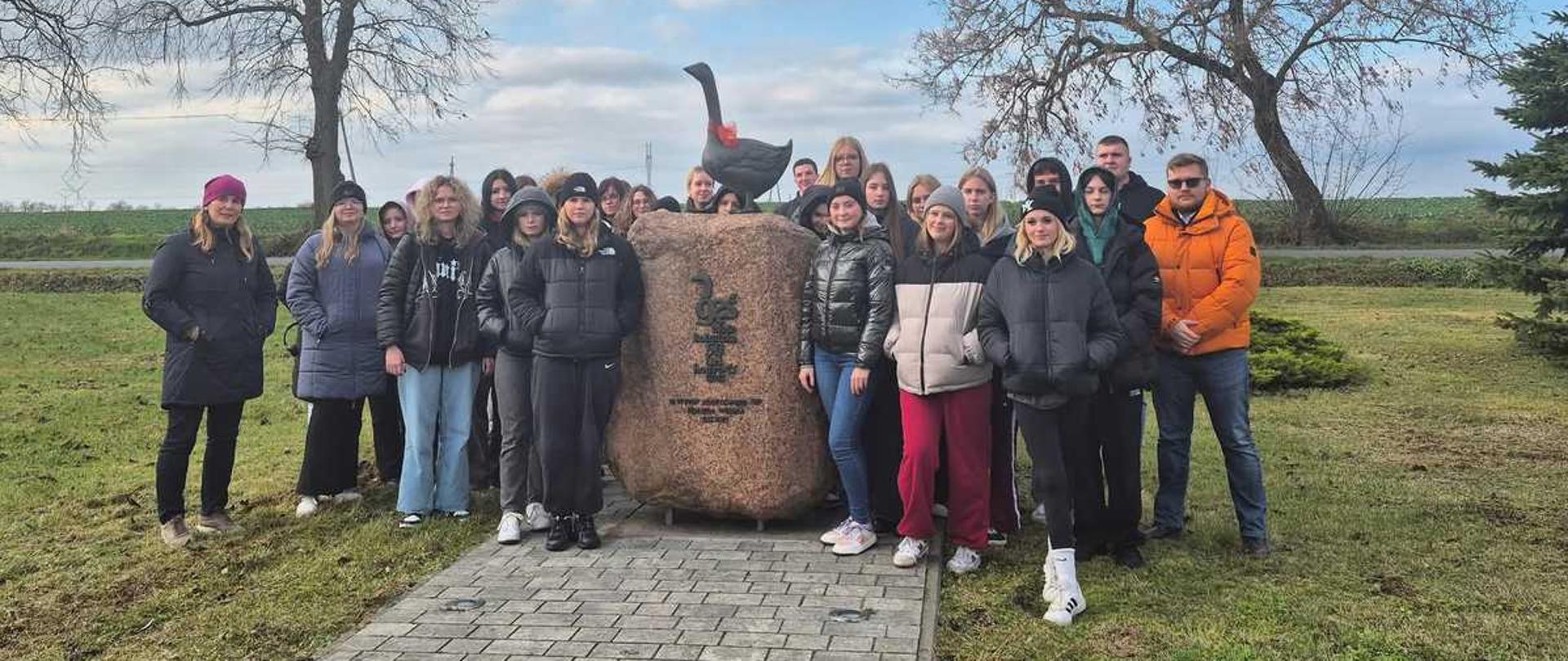 Zdjęcie przedstawia dużą grupę osób pozujących do wspólnego zdjęcia na zewnątrz, przed kamiennym pomnikiem z rzeźbą gęsią na szczycie.