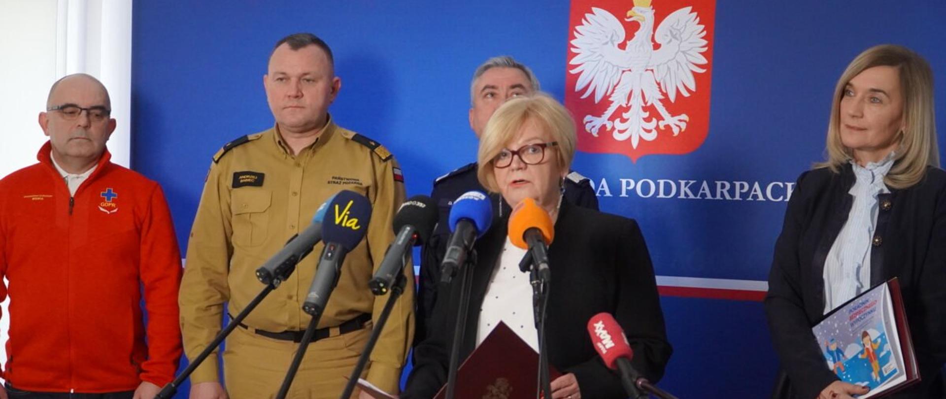 Zdjęcie zrobione w pomieszczeniu, podczas konferencji prasowej. Zdjęcie zrobione z dziennikarzy. Na tle banneru z napisem wojewoda podkarpacki stoją uczestnicy konferencji, wśród nich oficerowie Państwowej Straży Pożarnej i Policji. Przed mikrofonami stoi wojewoda podkarpacki i przemawia.