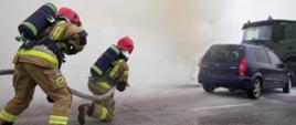 Strażacy w aparatach ochrony dróg oddechowych gaszą pożar samochodu osobowego i wojskowej cysterny, działając w gęstym dymie.