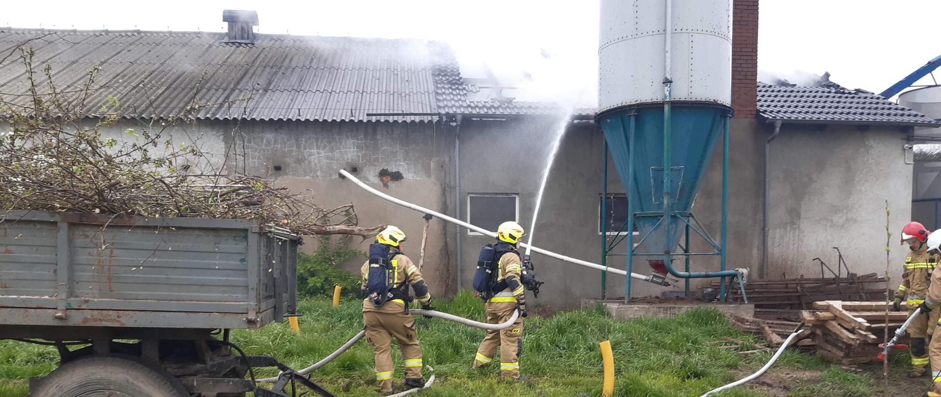 Pochmurno. Szary ciąg budynków. Przed budynkiem szara przyczepa. Palący się dach budynku z prawej strony. Dużo jasnego dymu. Dwóch strażaków w jasnych mundurach i białych hełmach z granatowymi butlami na plecach gasi dach budynku.