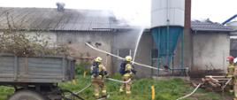 Pochmurno. Szary ciąg budynków. Przed budynkiem szara przyczepa. Palący się dach budynku z prawej strony. Dużo jasnego dymu. Dwóch strażaków w jasnych mundurach i białych hełmach z granatowymi butlami na plecach gasi dach budynku.