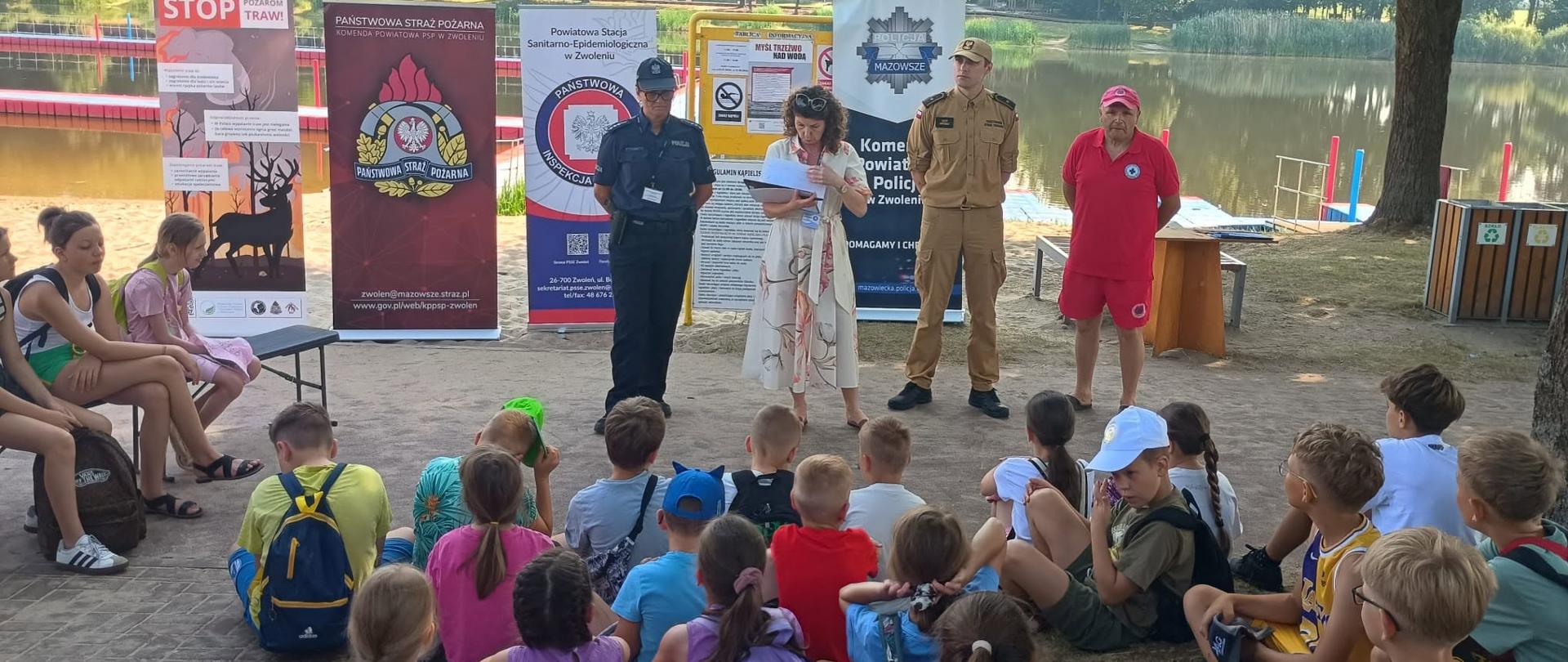 Zdjęcie grupowe wykonane na świeżym powietrzu nad brzegiem jeziora. Grupa dzieci siedzi na trawie, słuchając czworga dorosłych stojących przed nimi. Dorośli to: kobieta w kwiecistej sukience (czytająca kartkę), funkcjonariuszka policji w mundurze, funkcjonariusz straży pożarnej w mundurze polowym oraz ratownik w czerwonej koszulce i spodenkach. W tle, za prelegentami, stoją cztery duże banery reklamowe: jeden z napisem "STOP POŻAROM TRAW!", drugi z logiem Państwowej Straży Pożarnej, trzeci z logiem Inspekcji Sanitarnej i czwarty z logiem Policji. Jezioro z pomostem i drzewami widoczne jest w tle po lewej stronie.