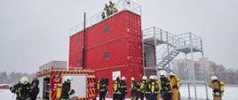 Obok trenażera stoi wóz strażacki oraz grupa strażaków i ratowników w kombinezonach i hełmach; na szczycie trenażera stoi trzech mężczyzn