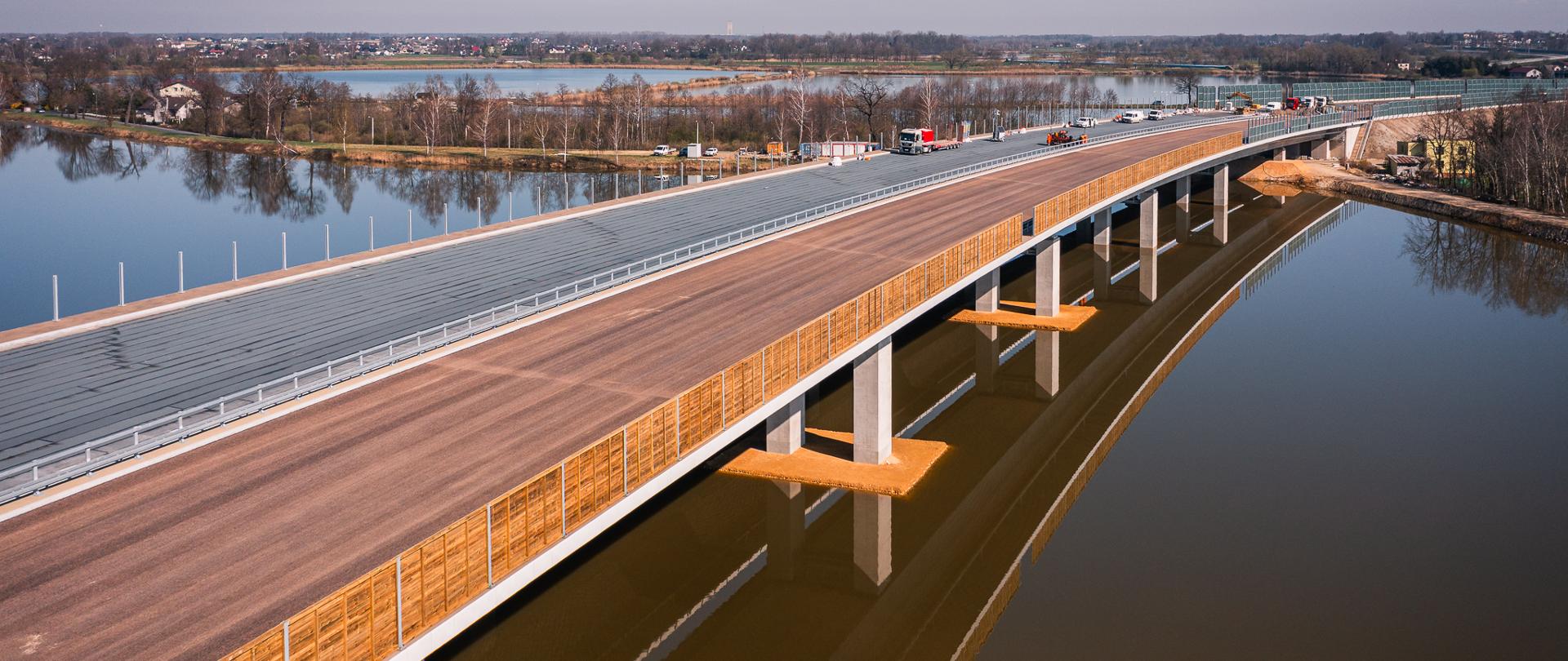 Na zdjęciu widoczny jest nowoczesny most wielopasmowy rozciągający się nad zbiornikiem wodnym. Konstrukcja wsparta jest na solidnych, betonowych filarach. Po moście poruszają się różne pojazdy, w tym samochody osobowe i ciężarówki. Powierzchnia wody poniżej działa jak lustro, odbijając sylwetkę mostu. W tle rozciąga się krajobraz z drzewami i zabudowaniami, a nad całością rozpościera się bezchmurne niebo.