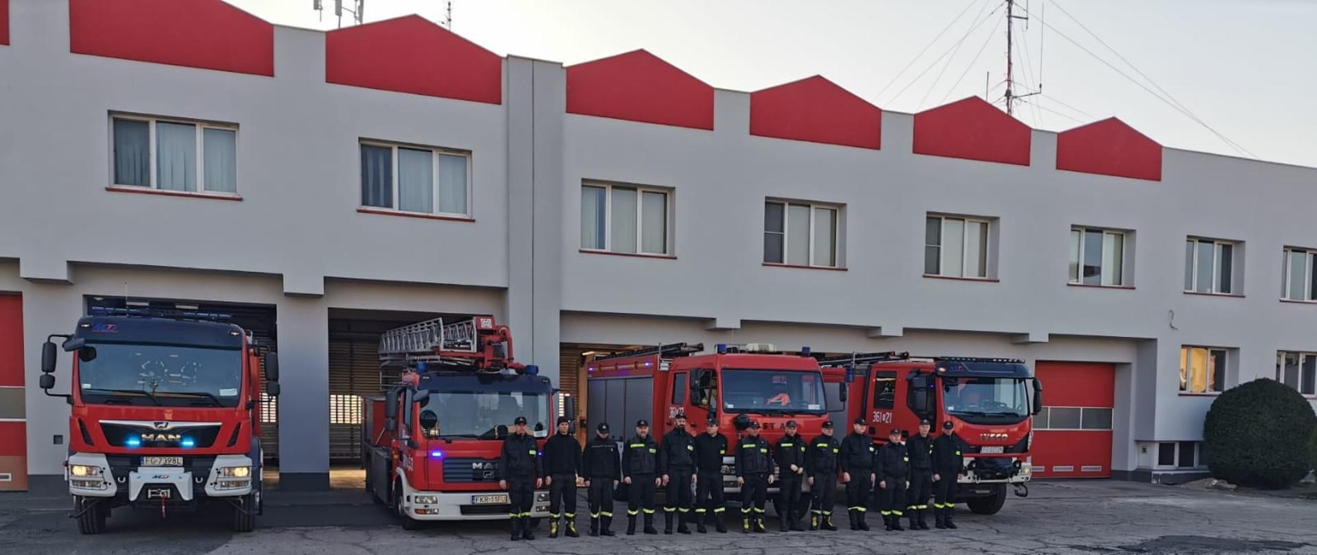 Na fotografii widoczni są strażacy oddający hołd dla strażaków z Ukrainy. Strażacy znajdują się przed jednostką na tle samochodów pożarniczych. Ubrani są w czarne mundury.