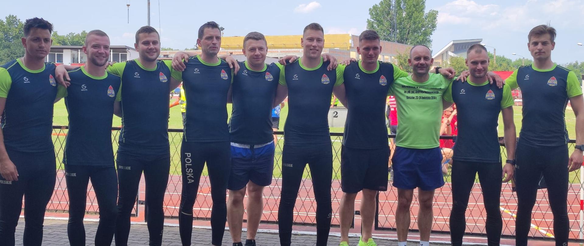 Zdjęcie grupowe drużyny strażaków z województwa opolskiego na XXXIX Międzynarodowych Mistrzostwach Polski w Sporcie Pożarniczym, które odbyły się w Centralnej Szkole Państwowej Straży Pożarnej w Częstochowie. W tle widoczny stadion z bieżnią. Strażacy w strojach sportowych.