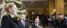 Zdjęcie z boku, minister Nowacka stoi i mówi do mikrofonu na stojaku, za nią choinka, dalej korytarz i schody.
