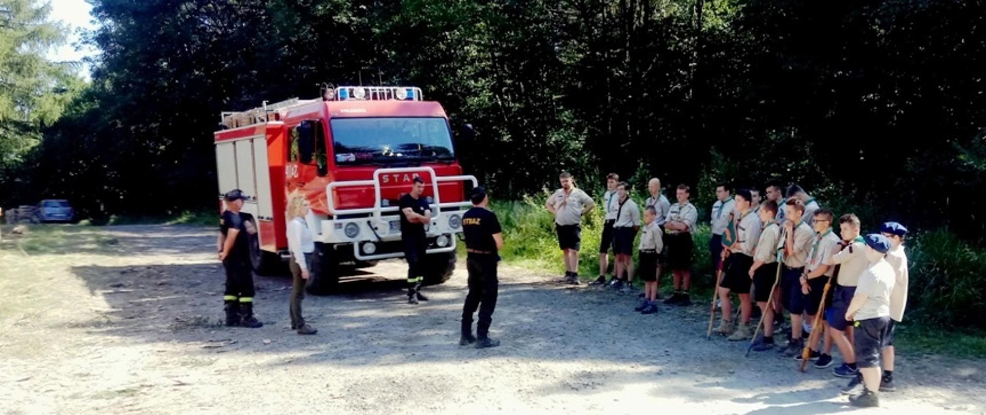 Zdjęcie przedstawia harcerzy na zbiórce ze strażakami w Jabłonicy Ruskiej. Na pierwszym planie kilkunastu harcerzy w szarych mundurach i kilku umundurowanych na czarno strażaków, przy czerwonym samochodzie ratowniczo-gaśniczym. Tło stanowi gęsty, wysoki, zielony las i jasne niebo. 