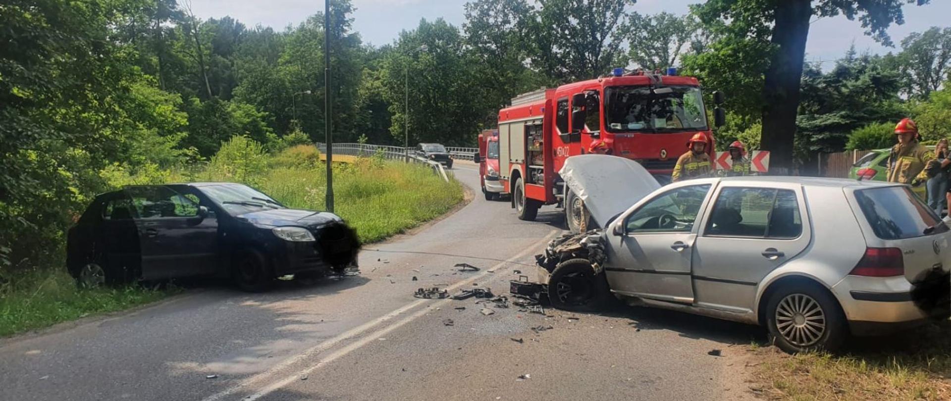 Na drodze i poboczu znajdują się samochody osobowe po zderzeniu czołowo-bocznym. Na jezdni znajdują się uszkodzone elementy pojazdów. Droga jest zablokowana.