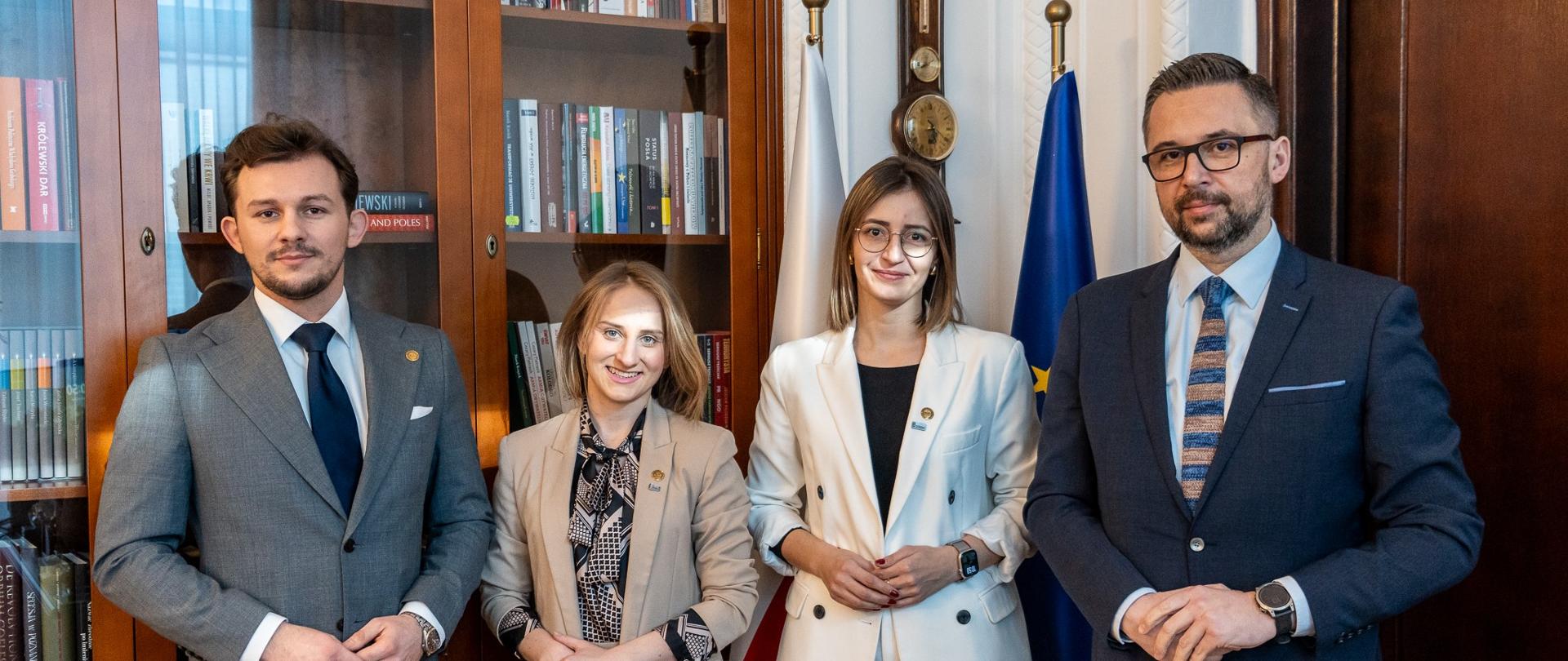 W małej sali stoi minister Kulasek i troje młodych ludzi, za nimi przeszklone szafy z książkami i pod ścianą flagi Polski i UE.