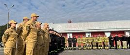 Strażacy stoją obok siebie za nimi znajduje się budynek służby ratowniczej.