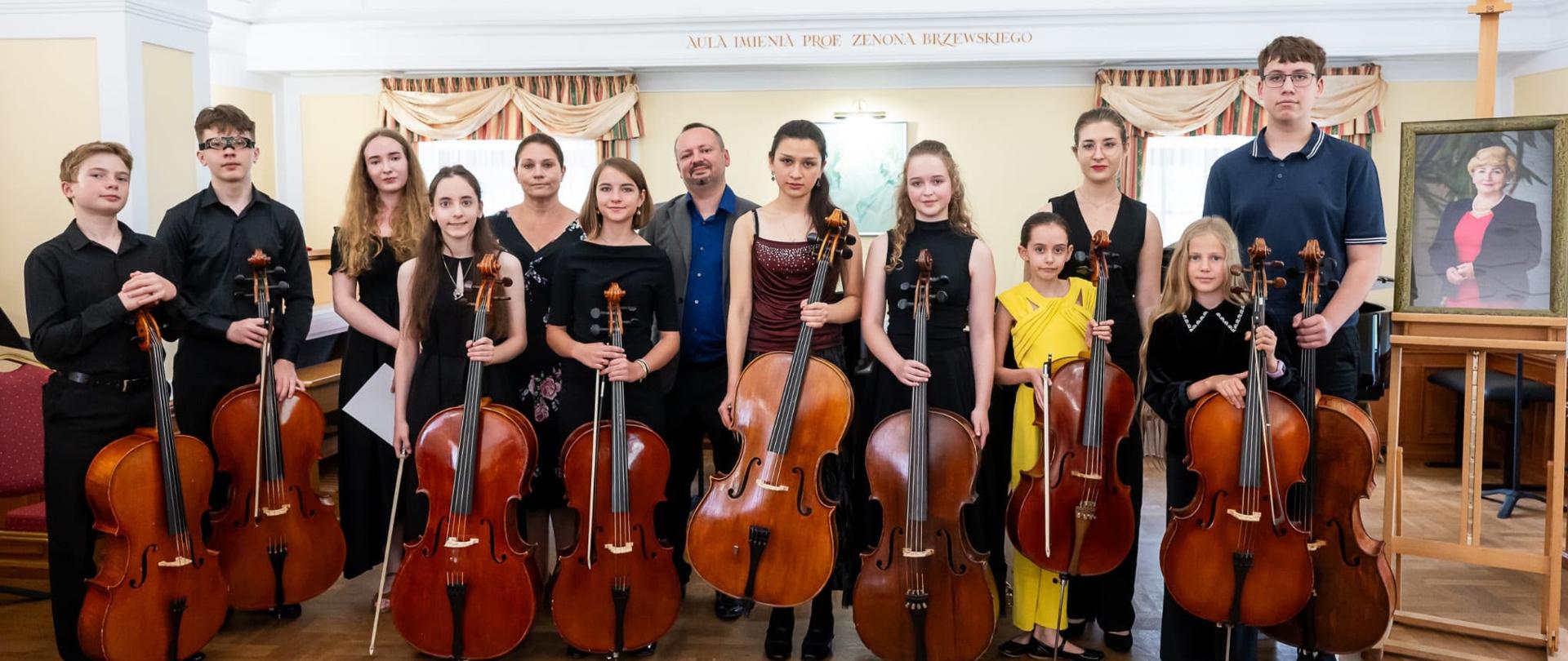 Zdjęcie grupowe osób w sali muzycznej, wszyscy trzymają wiolonczelę