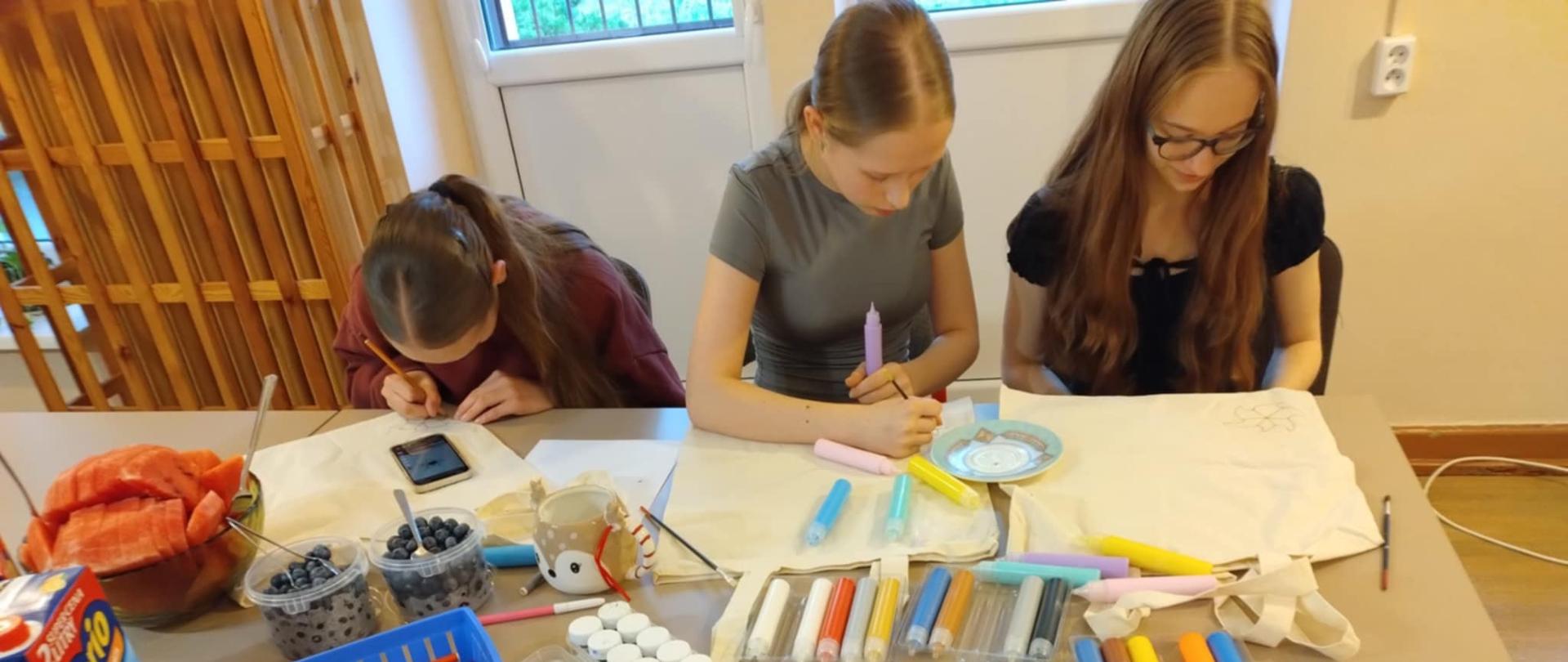 Trzy dziewczynki siedzące przy stole, zajęte pracami plastycznymi.