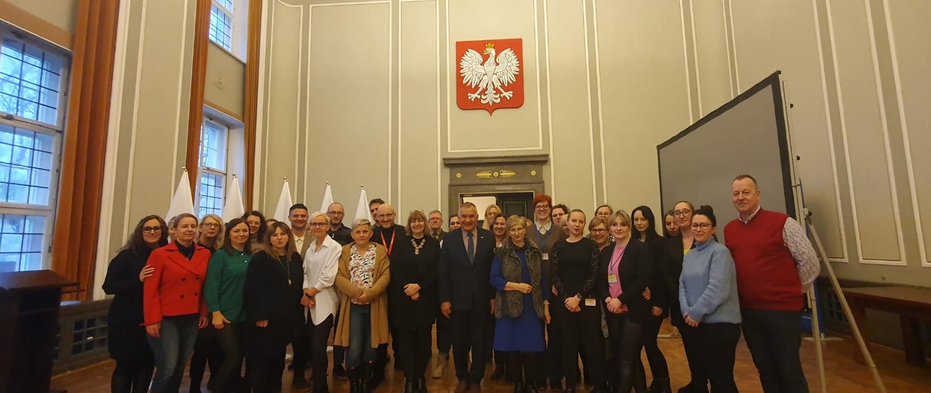 Kilkanaście osób stoi na tle Godła Polski. Nad głowami świeci okazały Żyrandol. 