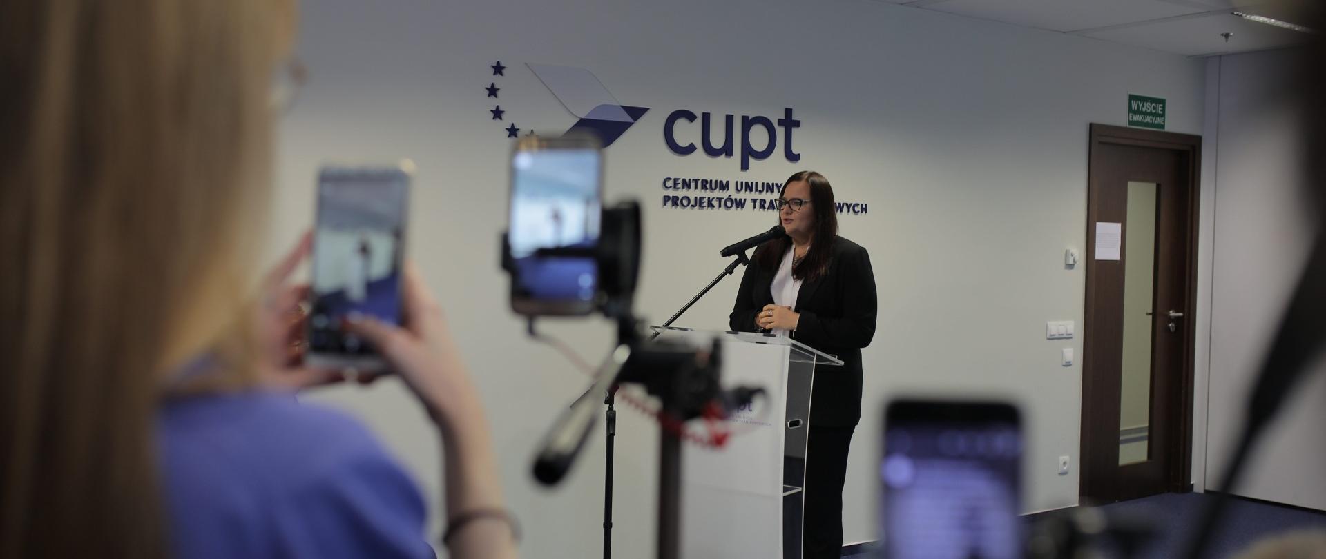 Minister Jarosińska-Jedynak stoi przy mównicy, mówi do mikrofonu. Za nią na ścianie wisi napis Centrum unijnych Projektów Transportowych. 