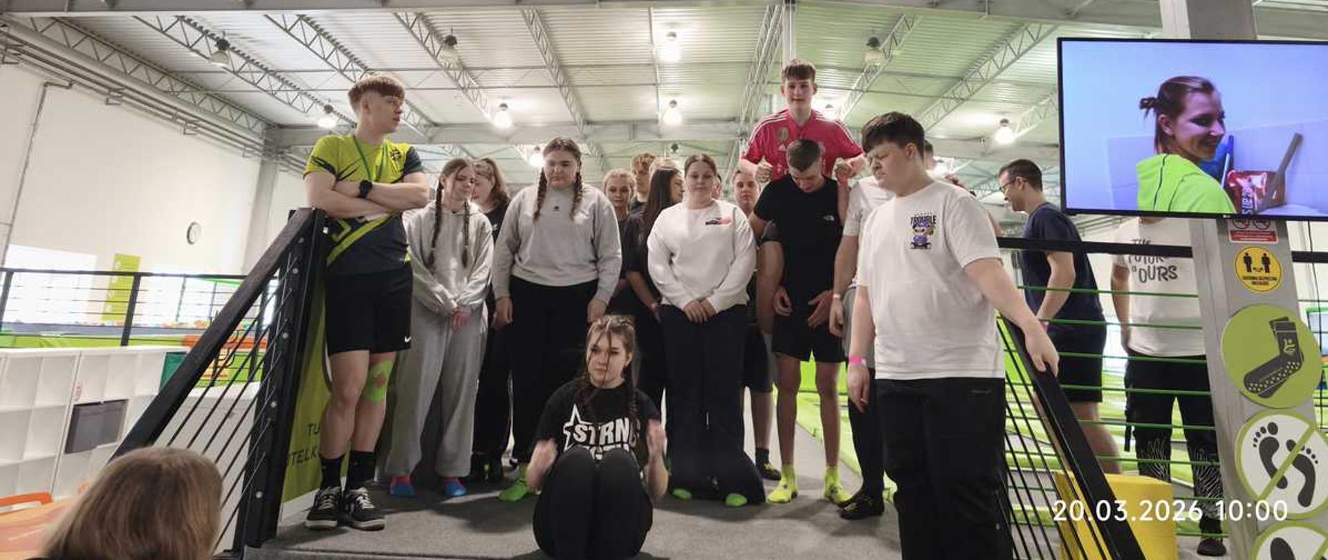 To zdjęcie przedstawia grupę młodzieży przebywającą w parku trampolin Jump Arena w Toruniu, co sugerują widoczne na miejscu oznaczenia.
Zdjęcie zostało wykonane 20 marca 2026 roku o godzinie 10:00.
Miejsce to jest popularną przestrzenią rekreacyjną przeznaczoną do aktywnego spędzania czasu, organizowania wycieczek integracyjnych oraz zajęć sportowych.
Na monitorze widocznym po prawej stronie wyświetlany jest materiał wideo z tego samego wydarzenia, co wskazuje na profesjonalną obsługę wizualną wizyty.
Uczestnicy spotkania znajdują się na specjalnej platformie, która stanowi punkt widokowy lub miejsce zbiórki przed rozpoczęciem aktywności na trampolinach.