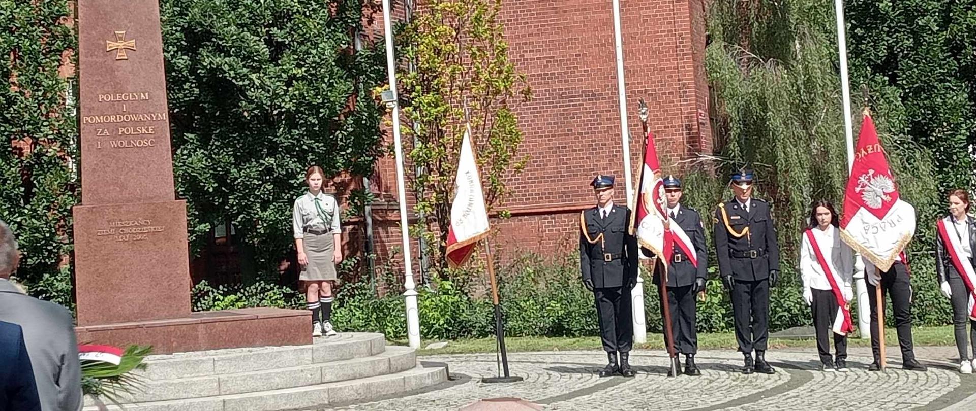 Zdjęcie przedstawia pomnik i stojące przy nim poczty sztandarowe podczas uroczystości 84. rocznicy wybuchu II wojny światowej.
W tle budynki i drzewa.
