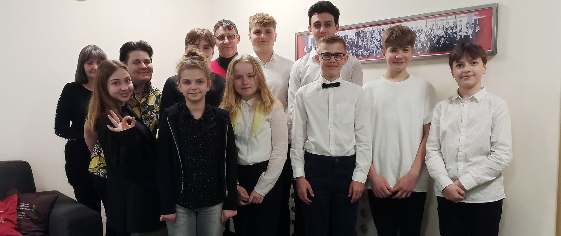 Na zdjęciu grupa uśmiechniętych uczniów i nauczycieli, 12 osób, stoją na tle białej ściany, jedna z dziewczynka pokazuje prawą ręką jakiś symbol, z prawej strony na ścianie wisi podłużny, prostokątny obraz o nieokreślonej treści. 