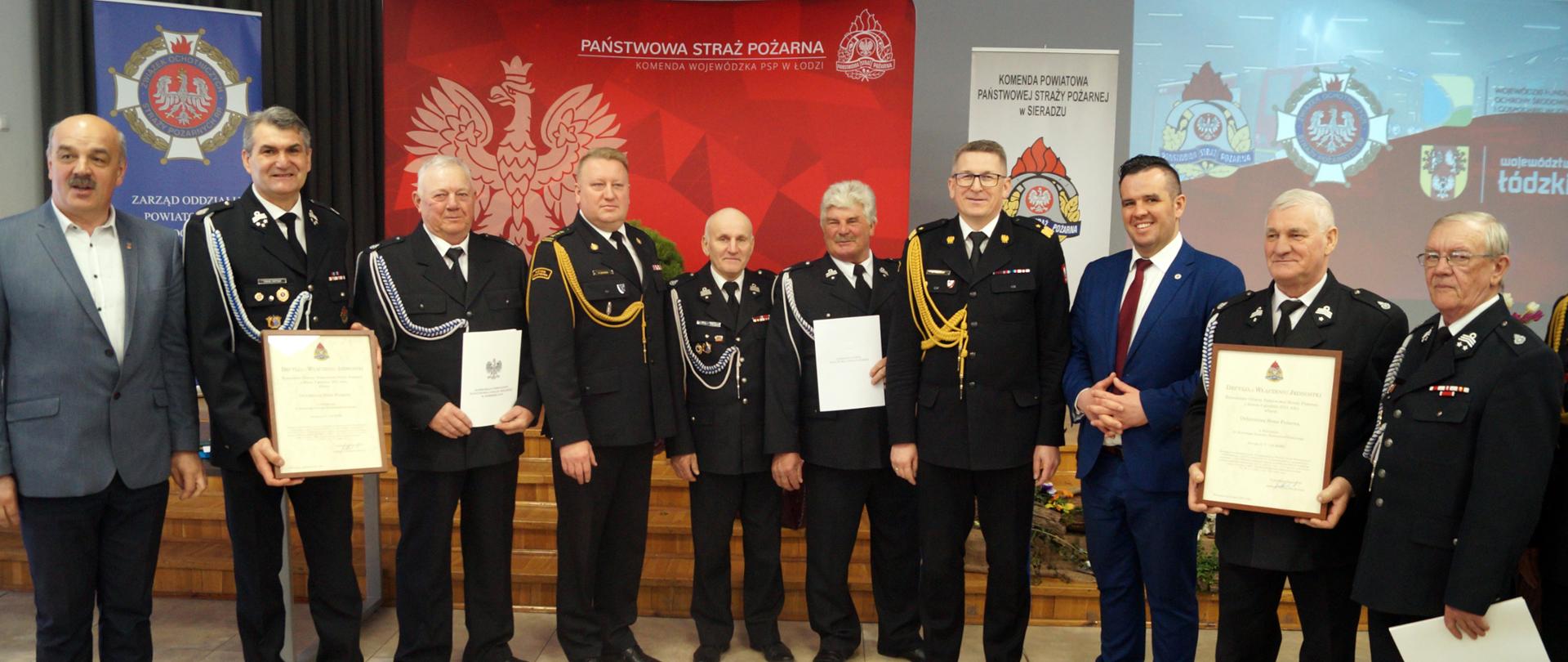 Na zdjęciu znajduje się doradca Prezydenta RP, Zastępca Komendanta Głównego nadbryg. Krzysztof Hejduk, razem z druhami OSP którzy trzymają akty włączenia do KSRG i decyzje o przyznaniu świadczenia ratowniczego. W tle banery PSP. 