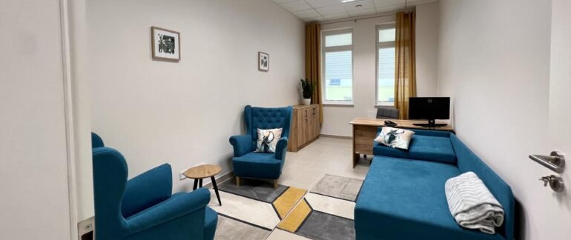 Wnętrze jednego z pomieszczeń Środowiskowego Domu Samopomocy w Rzeszowie przy ulicy Litewskiej - w środku dywan w geometryczne wzory, dwa niebieskie fotele, okrągły stolik, z prawej strony niebieski tapczan oraz biurko.