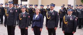 Zdjęcie przedstawia nagrodzonych medalem stających w oczekiwaniu na wręczenie medalu. w tle zgromadzeni strażacy.