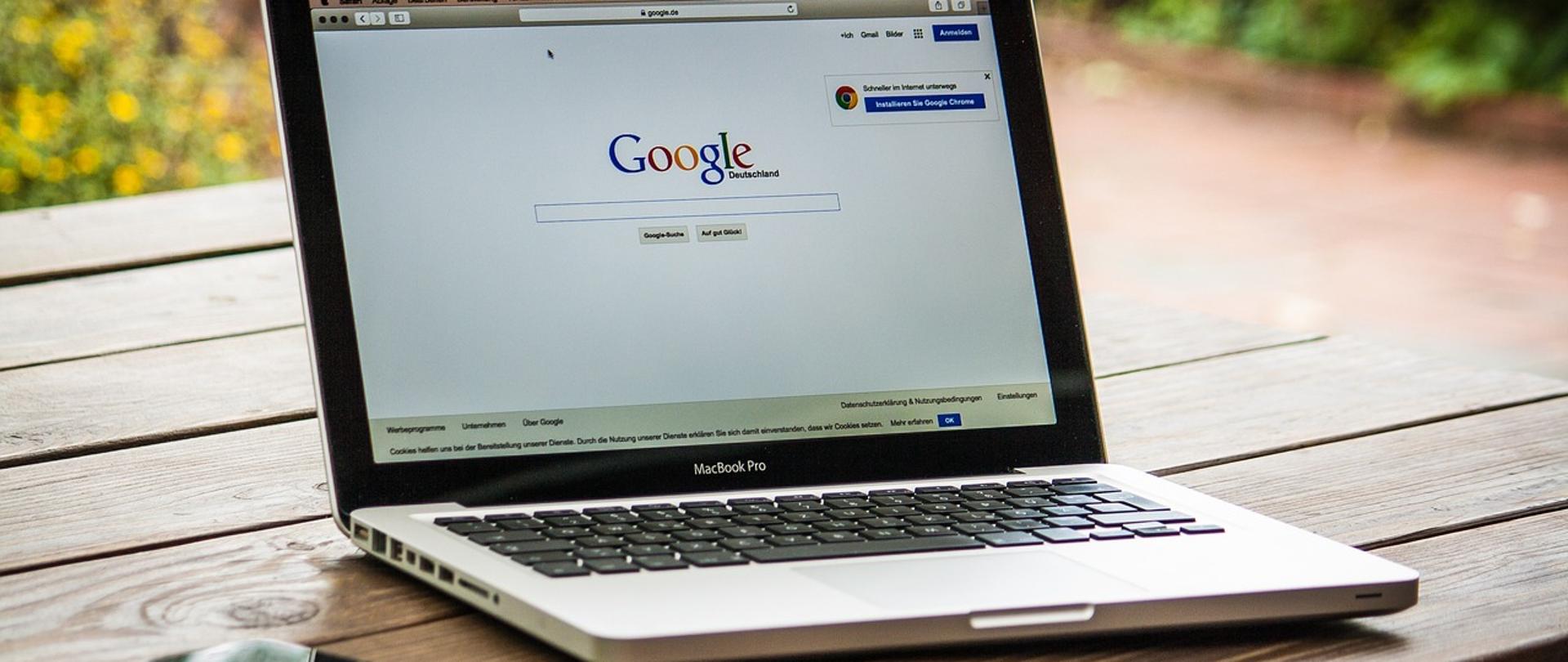 Zdjęcie laptopa na drewnianym stole z otworzonym oknem wyszukiwarki Google. Po lewej stronie smartphone