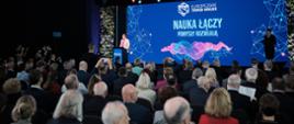 Zdjęcie od tyłu wypełnionej ludźmi sali, z przodu za mównicą stoi wiceminister Zioło-Pużuk i mówi do mikrofonu, za nią wielki ekran z napisem na niebieskim tle Nauka łączy - pomysły rozwijają.